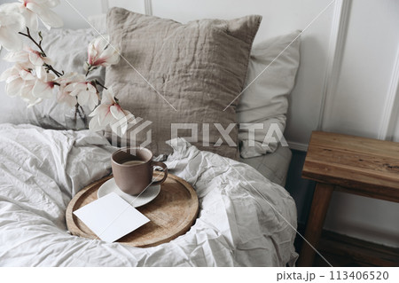 Breakfast in bed concept. Cup of coffee on wooden tray. Blank greeting card, invitation mockup. Bedroom view. Blurred beige linen pillows, blanket. Blooming magnolia branches in vase. Top view. Breakfast in bed concept. Cup of coffee on wooden tray. Blank greeting card, invitation mockup. Bedroom view. Blurred beige linen pillows, blanket. Blooming magnolia branches in vase. Top view. 113406520