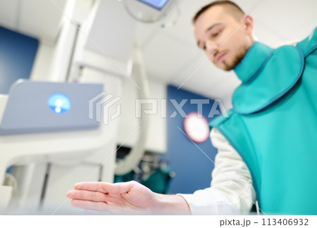 Young man having x-ray shot of broken hand in x-ray room in modern clinic. Patient wearing in protect lead apron Young man having x-ray shot of broken hand in x-ray room in modern clinic. Patient wearing in protect lead apron 113406932
