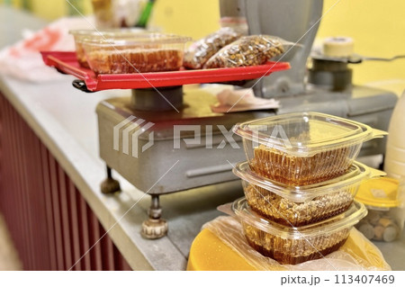 Honeycombs with honey in transparent plastic containers on the counter for sale lie next to the scales. Wax structures made of honey in the food market Honeycombs with honey in transparent plastic containers on the counter for sale lie next to the scales. Wax structures made of honey in the food market 113407469