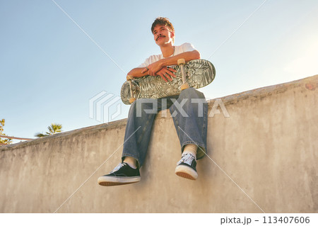 Smiling young male skateboarder holding skateboard sitting on ramp in skate park Smiling young male skateboarder holding skateboard sitting on ramp in skate park 113407606