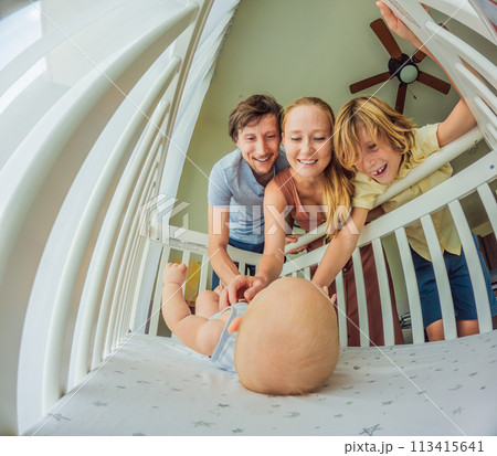 Big family with two kids in white bedroom. Parents and kids standing at crib of newborn baby boy. Boy meeting new sibling. Nursery for infant Big family with two kids in white bedroom. Parents and kids standing at crib of newborn baby boy. Boy meeting new sibling. Nursery for infant 113415641