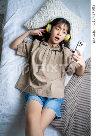 ヘッドフォンをしてベッドでスマホを操作する女の子 113417905