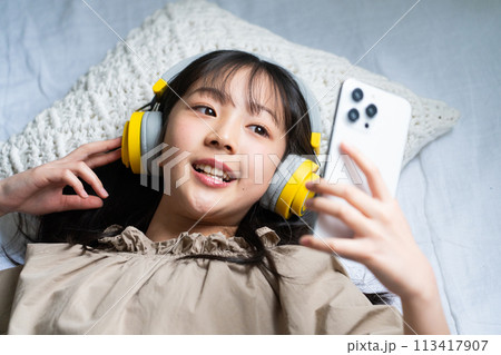 ヘッドフォンをしてベッドでスマホを操作する女の子 113417907