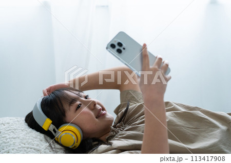 ヘッドフォンをしてベッドでスマホを操作する女の子 113417908