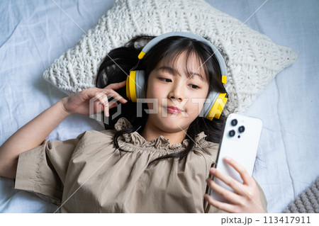 ヘッドフォンをしてベッドでスマホを操作する女の子 ヘッドフォンをしてベッドでスマホを操作する女の子 113417911