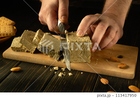 Close-up of a man hands using a knife to cut halva on a cutting board. Sweet and tasty breakfast concept with sunflower halva and tea at home 113417950
