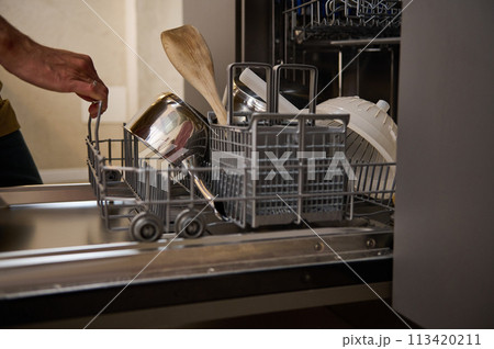 Handsome man chef-cook in beige chef's apron, emptying out, loading or unloading, taking clean dishes out of a modern automatic dishwasher at minimalist home kitchen interior. People. Household chores 113420211