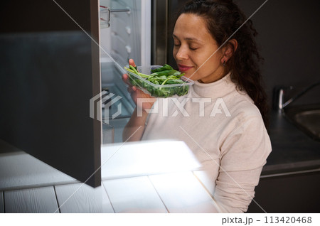 Charming multi ethnic woman holding box with fresh arugula leaves, smelling it, standing by open fridge in the home kitchen. 113420468