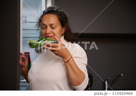 Multi ethnic woman pulls out a recyclable cardboard crate with ecologically friendly organic green spinach leaves from the refrigerator, enjoying the aroma of fresh organic herbs while cooking salad 113420469