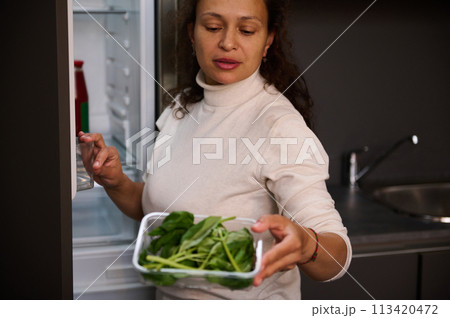 Pretty woman standing by the fridge, pulling out a recyclable paper box with ecologically friendly organic greens and herbs, spinach leaves from the refrigerator. cooking healthy salad Pretty woman standing by the fridge, pulling out a recyclable paper box with ecologically friendly organic greens and herbs, spinach leaves from the refrigerator. cooking healthy salad 113420472