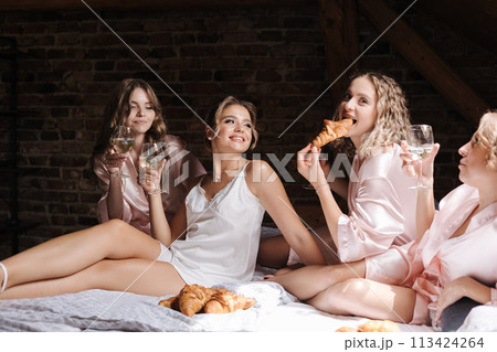 Portrait of four females sitting on bed and eating croissants. Bridesmaids and bride wears silk white robe. Woman drinks champagne 113424264