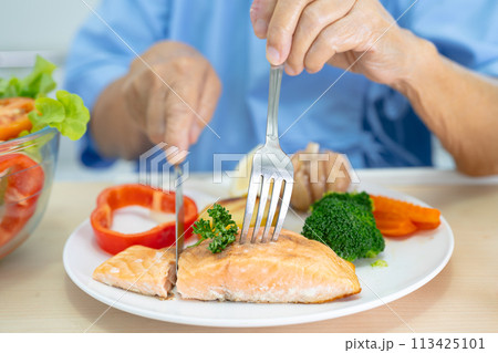 Asian senior woman patient eating Salmon steak breakfast with vegetable healthy food while sitting and hungry on bed in hospital. Asian senior woman patient eating Salmon steak breakfast with vegetable healthy food while sitting and hungry on bed in hospital. 113425101