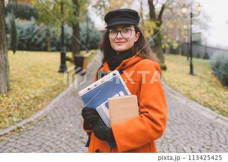 A woman wearing an orange coat and hat is holding a stack of books 113425425
