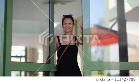 Beautiful smiling young woman opens glass door Beautiful smiling young woman opens glass door 113427789