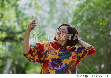 Cheerful young lady with glasses capturing a selfie in a lush green park. Young entrepreneur with digital devices capturing a selfie, staying connected in an urban green space Cheerful young lady with glasses capturing a selfie in a lush green park. Young entrepreneur with digital devices capturing a selfie, staying connected in an urban green space 113427951