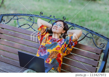 Asian freelancer with laptop enjoying relaxing on bench in park in summer Freelance lifestyle portrayed by a woman engaging with technology in a peaceful park setting 113427972