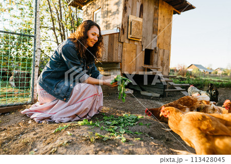 Happy middle aged woman on a private farm feeding chickens. Joyful farmer woman caring for her bird in her backyard in a rustic style, demonstrating an eco-friendly lifestyle. Sustainable lifestyle 113428045