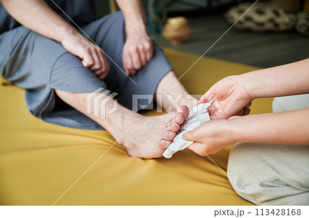 Close up of woman masseuse cleaning by soft wet tissue male client's feet before Thai massage on the mat. Cropped view. Close up of woman masseuse cleaning by soft wet tissue male client's feet before Thai massage on the mat. Cropped view. 113428168