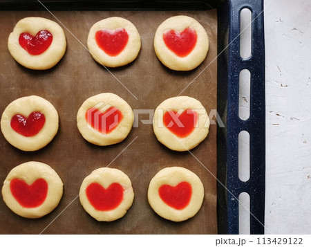 Round shortbread cookies with a red heart decor, which is filled with strawberries with sugar and strawberry jam, lying on a craft parchment on a blue baking sheet standing on a white wooden table 113429122
