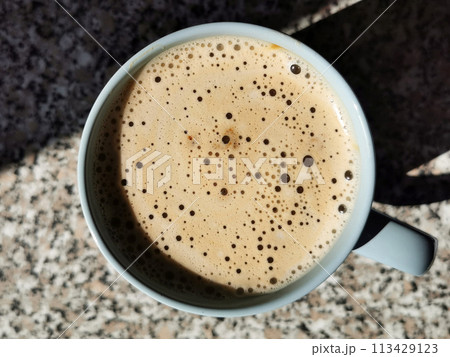 Top view of a blue coffee mug with beige milk foam with bubbles of different sizes, which stands on a mottled chipboard countertop with a stone texture 113429123
