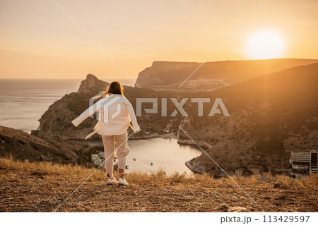 A woman stands on a hill overlooking a body of water. The sun is setting in the background, casting a warm glow over the scene. The woman is enjoying the view and taking in the beauty of the landscape 113429597
