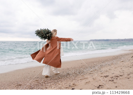 Blond woman Christmas tree sea. Christmas portrait of a happy woman walking along the beach and holding a Christmas tree on her shoulder. She is wearing a brown coat and a white suit. 113429668