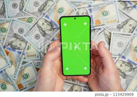 Hands holding smartphone with green screen on pile of dollar banknotes, top view. Concept of online earnings 113429909