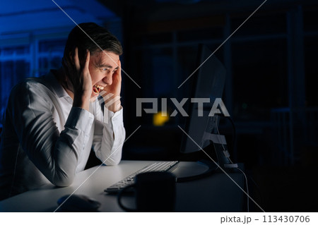 Angry office worker male working late at night and looking at computer while screaming alone. Furious man working at workplace night crazy and mad shouting, yelling with aggressive expression 113430706