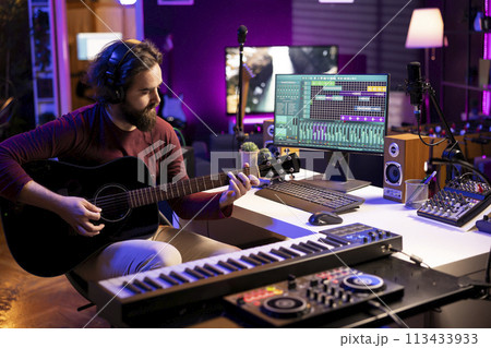 Young singer playing acoustic guitar and creating music with electronic tools in home studio, using daw software interface on pc. Sound producer recording tunes and mixing sounds on stereo console. 113433933