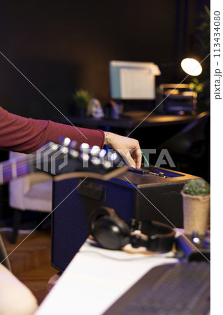 Sound producer adjusting volume levels on home studio amplifier before recording guitar tunes for mixing and mastering session. Male composer creating a new song with stereo gear. Close up. Sound producer adjusting volume levels on home studio amplifier before recording guitar tunes for mixing and mastering session. Male composer creating a new song with stereo gear. Close up. 113434080
