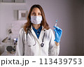 A young adult female doctor in a medical mask and white coat holds a syringe in her hands and looks at the camera 113439753