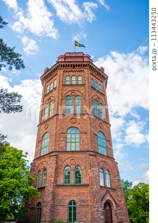 Bredablick Tower stands tall against a blue, cloud-speckled sky at Skansen in Stockholm, Sweden, showcasing its brick architecture and Swedish flag. Bredablick Tower stands tall against a blue, cloud-speckled sky at Skansen in Stockholm, Sweden, showcasing its brick architecture and Swedish flag. 113443250