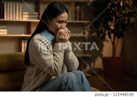 Young Asian Woman Blowing Her Nose in a Cozy Home Setting During Evening Young Asian Woman Blowing Her Nose in a Cozy Home Setting During Evening 113446530