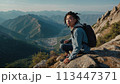 A young happy Chinese man with a tourist backpack stands on top of a rock in the mountains, travels and enjoys a beautiful view of the mountains, sky or nature. Untouched nature. Tourism and travel 113447371