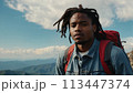 Young positive African American man with a tourist backpack stands on top of a rock in the mountains, travels and enjoys a beautiful view of the mountains, sky or nature. Untouched nature. Tourism and 113447374