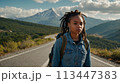 Young positive African American woman with a tourist backpack stands on top of a rock in the mountains, travels and enjoys a beautiful view of the mountains, sky or nature. Untouched nature. Tourism 113447383