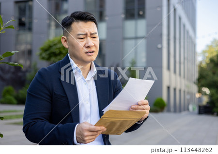 Upset and disappointed Asian businessman sitting outside the office and reading a received letter with bad news. 113448262
