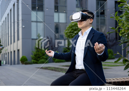 A young man in a business suit sits on a bench outside a company building in a lotus position, meditating in a virtual mask online. A young man in a business suit sits on a bench outside a company building in a lotus position, meditating in a virtual mask online. 113448264