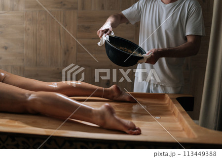 Cropped shot of unrecognizable practitioner male pouring heated sand from bowl onto massage table for scrubbing treatment to female client. Preparation to serenity of recreation at spa salon. Cropped shot of unrecognizable practitioner male pouring heated sand from bowl onto massage table for scrubbing treatment to female client. Preparation to serenity of recreation at spa salon. 113449388