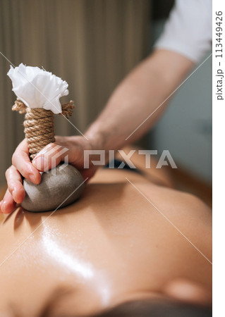 Cropped vertical shot of Ayurveda Massage therapist pressing herbal bolus bags onto female skin. Masseur gently compresses herb bag on woman body. Hot herbal ball spa massage body treatment. 113449426