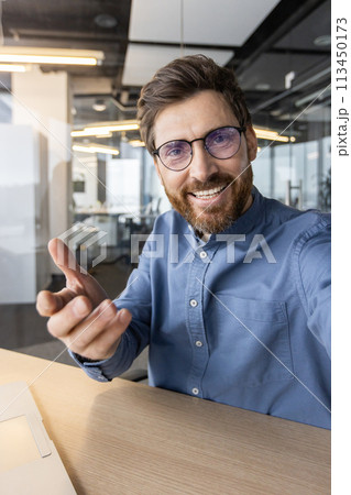 Happy bearded man in a blue shirt reaching towards the camera, offering a greeting with a warm, friendly smile in an office setting. 113450173