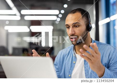Serious young hispanic man in headset sitting at desk in office and talking on video call. Close-up photo. 113450204