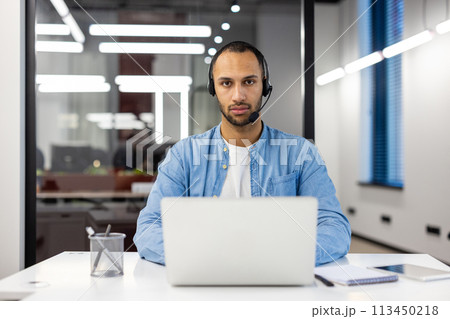 Portrait of a serious young hispanic male specialist wearing a headset, sitting at a desk in a busy office, working on a laptop, looking intently at the camera. 113450218
