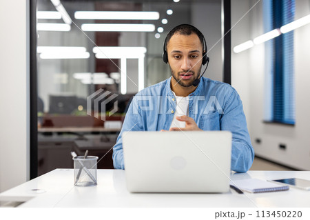 Latin American man in a headset sits in the office in front of a laptop and communicates on a video call. 113450220
