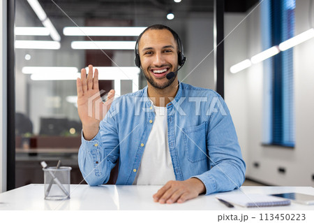 Smiling young African American man in headset sitting in office in front of camera, talking and waving hand to camera. 113450223