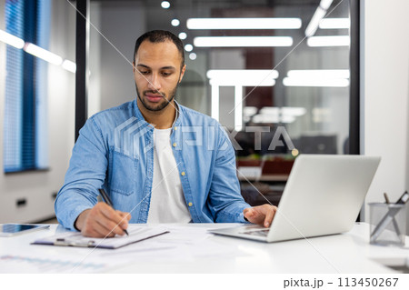 Focused Indian businessman at his desk in a well-lit modern office, working on a laptop and writing notes. Focused Indian businessman at his desk in a well-lit modern office, working on a laptop and writing notes. 113450267
