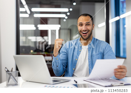 Successful Indian businessman fist-pumping in celebration at his modern office workspace, feeling excited and happy. 113450283