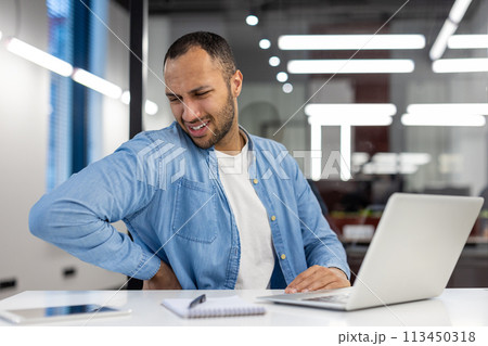 An Indian man in a living room setting appears to suffer from back pain while working on his laptop, portraying common workplace discomfort. 113450318