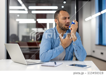 A man of Indian descent is in an office setting, using an asthma inhaler, showing signs of respiratory distress. 113450319