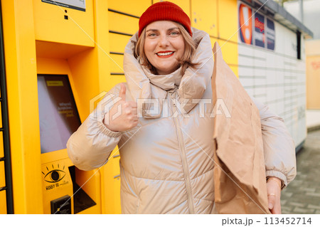 Happy smiling woman client with box at outdoor automated parcel machine choosing operation on touch screen by smart phone Happy smiling woman client with box at outdoor automated parcel machine choosing operation on touch screen by smart phone 113452714